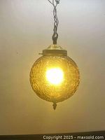 Front view of the amber swag lamp illuminated, showing the embossed floral swirl pattern on the glass globe and hanging chain.