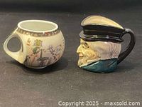 Two ceramic mugs shown side by side on a wooden surface. Left mug has ancient Egyptian design, right mug is character face shape.