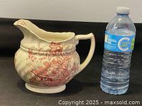 Side view of cream-colored ceramic Royal Doulton pitcher with red floral pattern and wide spout, shown beside water bottle for scale.