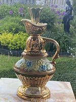 Front view of a vintage decanter with ornate gold detailing and pastel floral accents on the base, positioned outdoors against a garden backdrop.