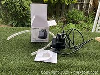 Photo showing Aquamcom bottom submersible pump, power cord, two clear plastic tubes, manual, and box on grass surface.