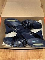 Pair of navy blue Nike Air Max Tremble sneakers seen inside an open shoebox on a wooden floor, showing black horizontal straps, blue upper, and a visible air sole.