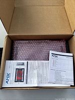 Opened box showing one panel wrapped in pink bubble wrap alongside printed AMX safety instructions and manual.