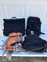 Four bags placed on table: a black briefcase, black tactical backpack, orange Gregory hiking backpack with visible wear, and a black duffel bag