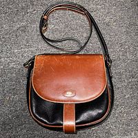 Full view of Bally small leather crossbody shoulder bag, black textured body with brown leather flap and strap laid flat on carpet