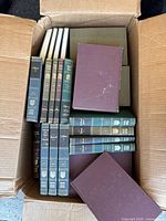 Top view of the open box showing 54 volumes in green and brown hardcovers with gold and white spine text, arranged lying flat and standing.