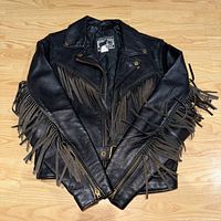 Full front view of black leather motorcycle jacket with fringe detail on sleeves and chest, lying flat on wooden floor.