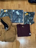 All five shirts and one brown hooded jacket shown on wood floor, shirts three new in packaging, one maroon pocket shirt visible, jacket unzipped showing black lining