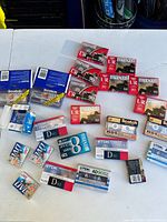 Lot of cassette tapes and camcorder tapes on a white table including Maxell, Scotch, TDK, and JVC brands.