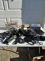 Full view of the lot showing vintage General Electric radio cassette player, remote controls, headphones, keyboards, power adapters and other electronic accessories spread on a table outdoors.