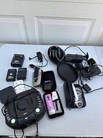 Dark table with a black portable CD player with control buttons center, over-ear headphones and carrying case, two black rectangular battery packs labeled 'Radio Slave II', power adapter, a black small pouch, pink and white personal care device in mesh pouch, and assorted cables scattered on the table.