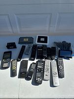 Various remote controls and two calculator devices laid out on a white surface with cables and two splitter box devices.