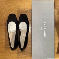 Top view of Everlane black leather ballet flats next to original Everlane Italy branded box, flat on wooden surface.