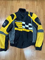 Front view of Honda branded motorbike jacket in black, yellow and gray with visible logos and patches.