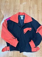 Front view of Kansai O2 wool jacket laid flat showing color-block black and orange body with button front and flap pocket detail.