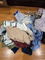 Large pile of assorted men's shirts on wood floor, showing variety of fabrics and colors including striped and solid patterns