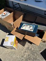 Overview of multiple cardboard boxes containing toys, household items, skateboard deck partially visible with Hot Wheels logo.