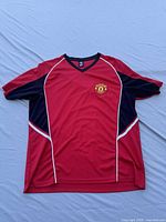Full front view of the red Manchester United soccer jersey with black side panels and white piping accents laid flat on a light blue surface.