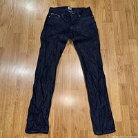 Full front view of blue raw selvedge denim jeans laid flat on floor showing shape, color, and wear condition.