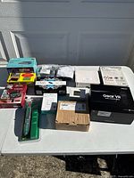 Wide one-table display of multiple new or sealed electronic items in original packaging set outdoors on a white folding table; includes networking devices, a VR headset box, and other assorted electronics.