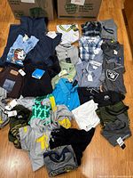 Wide view of the lot showing multiple clothing items including jacket, t-shirts, and pants with tags.
