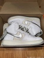 Pair of white Nike Air Force 1 High L/M 20Th sneakers with metallic silver swooshes in a brown box showing the side profile and ankle straps.