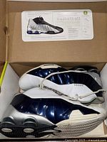 Pair of Nike Shox BB4 basketball sneakers inside the box showing top and side views with white, blue, and silver colors and Nike Shox technology visible on soles.