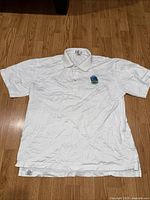 White men's polo shirt with Golden State Warriors embroidered logo on left chest, laid flat on wood floor.