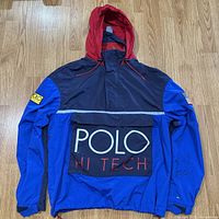 Full front view of Polo Ralph Lauren Hi Tech windbreaker jacket showing blue and navy color blocking, red hood, POLO HI TECH front pocket, and yellow Polo Hi Tech logo patch on left sleeve.
