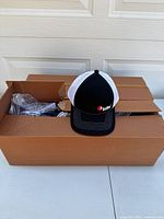 Open box showing RedMax hat on top with black front and white mesh back, embroidered logo on front.