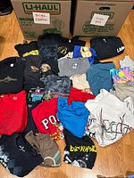 Overall lot showing variety of folded men's t-shirts alongside two U-Haul boxes labeled for sorting clothes and pants.