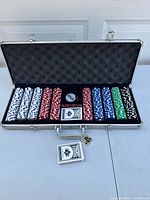 Open aluminum case showing poker chips organized by color in rows, two decks of playing cards, dealer button with key and red dice.