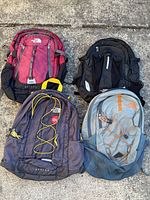 Four The North Face backpacks laid out on pavement showing different colors and styles: red/black, black, dark gray/yellow, gray/orange.