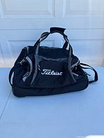 Front and side view of the black Titleist roller duffel bag showing carrying handles, front zippered compartment with embroidered Titleist logo.