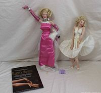 Two Marilyn Monroe Heirloom dolls side by side, one in pink dress with gloves and one in white pleated dress, accompanied by booklet