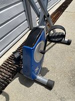 Close-up of the blue casing around the pedal mechanism, showing the foot pedal with strap and sturdy metal frame with black stabilizing feet.