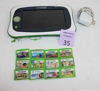 Front view of LeapPad Ultimate handheld console powered off, showing screen, buttons, attached strap, 11 green game cartridges laid out, and a white charging adapter.
