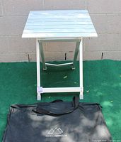 Aluminum camping table set up outdoors with green artificial grass and brick wall background, carrying bag placed in front.