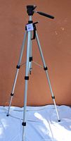 Full view of the silver tripod fully extended showing the three-section legs and the overall size, standing on a white cloth background against a plain wall.