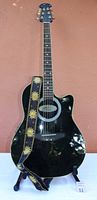 Full front view of black Ovation Applause AE700 guitar with sun-patterned strap on stand