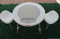 Front view of white Skip Hop play table with two white child chairs on each side on green outdoor carpet. The table has a circular grey removable center panel.