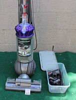 Dyson Animal Ball vacuum cleaner upright with purple cyclone and clear dust bin, plastic box with vacuum attachments, cleaner head on the ground