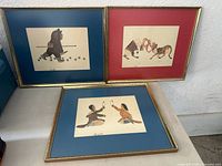 Three framed Inuit paintings arranged with two upright and one laid flat, showing varying mat colors and metallic frames.