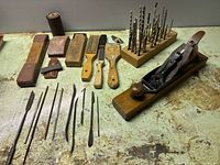 Wide shot showing assorted wood shaping/carving tools, files, scrapers, paint brushes, drill bits on wooden block, and a vintage wood plane on a work surface.