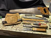 Photo showing antique woodworking tools including a wooden hand saw, bullnose rabbit planer, suede tool belt, chisels, slide tool, socket tool, and other hand tools arranged on a workbench.