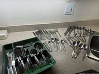 Flatware laid out on countertop and inside a green utensil organizer tray showing knives, forks, spoons, and serving pieces.