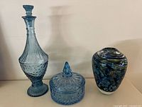 Three blue glass items on display: a tall carafe with stopper, a lidded candy dish, and a blue mottled blown glass vase.