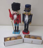 Two wooden nutcrackers standing side by side with six gold toned plastic apple ornaments in front, displayed in white boxes.
