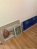 Three framed artworks leaned against wall: large signed hockey print, smaller Sheep Shearer print, wide blue replicate stained glass poster.