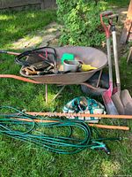 Overall view of the garden tools lot showing wheelbarrow loaded with tools, shovels leaning on the wheelbarrow, garden hose coiled on ground.
